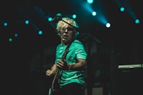 A musician with blond hair passionately plays an electric guitar on stage under vibrant blue lights. The atmosphere is energetic, and the musician appears to be engrossed in the performance.
