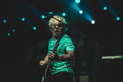 A musician with blond hair passionately plays an electric guitar on stage under vibrant blue lights. The atmosphere is energetic, and the musician appears to be engrossed in the performance.