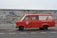 A red vintage van is parked on the street in front of a vibrantly painted graffiti wall. The wall features intricate, colorful designs with a mix of abstract shapes and detailed patterns. Words and motifs in various colors are interwoven into the mural. The van has branding on its side advertising a beverage company and displays German text.