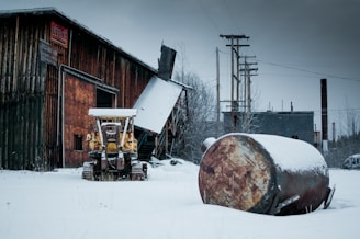 An abandoned industrial site covered in snow, featuring a rusted old bulldozer and a large metal tank lying on its side. The building has wooden panels and there are signs indicating its use as an office for hardware and supplies. Trees, power lines, and another industrial building are visible in the background under a gray, overcast sky.