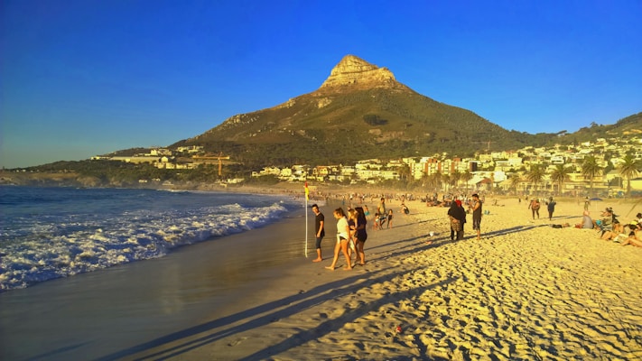 A sandy beach filled with people enjoying the sunny day. In the background, a prominent mountain with a distinct peak rises, surrounded by a lush green hillside. The beach is lined with white, modern buildings and palm trees. The ocean waves gently wash ashore under a clear blue sky.