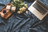 A laptop is placed on a dark blue checkered fabric, surrounded by a bouquet of light pink roses, a wooden tray holding a cup of coffee, and a pen. Additionally, a stylish wallet and a watch with a brown leather strap are on the fabric.