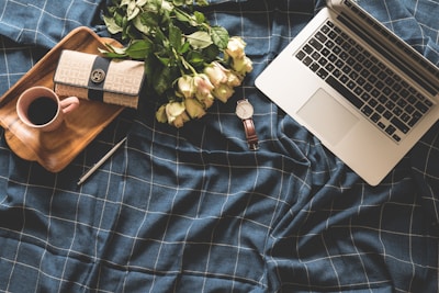 A laptop is placed on a dark blue checkered fabric, surrounded by a bouquet of light pink roses, a wooden tray holding a cup of coffee, and a pen. Additionally, a stylish wallet and a watch with a brown leather strap are on the fabric.