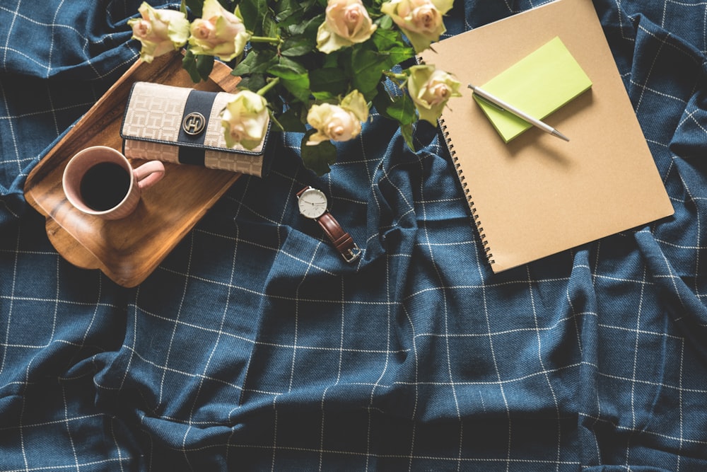 white rose flower bouquet beside brown note, round white analog watch with brown leather band and rectangular brown wooden serving tray on blue and white blanket close-up photo