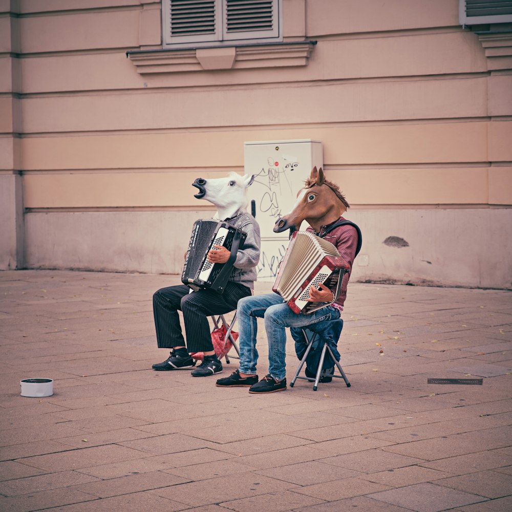 Zwei Personen mit Pferdeköpfen sitzen auf Klappstühlen, während sie Akkordeons neben einem braunen Betongebäude spielen