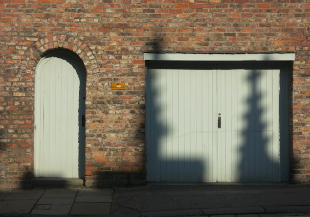 A red brick wall features an arched doorway with a wooden door on the left and a large double wooden garage door on the right. Shadows of tree branches are cast on the garage door. A small rectangular sign is mounted on the wall between the two doors. The ground in front of the doors is paved with stone slabs and concrete.