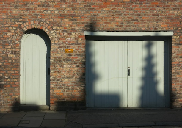 A red brick wall features an arched doorway with a wooden door on the left and a large double wooden garage door on the right. Shadows of tree branches are cast on the garage door. A small rectangular sign is mounted on the wall between the two doors. The ground in front of the doors is paved with stone slabs and concrete.