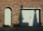 A red brick wall features an arched doorway with a wooden door on the left and a large double wooden garage door on the right. Shadows of tree branches are cast on the garage door. A small rectangular sign is mounted on the wall between the two doors. The ground in front of the doors is paved with stone slabs and concrete.
