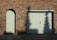 A red brick wall features an arched doorway with a wooden door on the left and a large double wooden garage door on the right. Shadows of tree branches are cast on the garage door. A small rectangular sign is mounted on the wall between the two doors. The ground in front of the doors is paved with stone slabs and concrete.