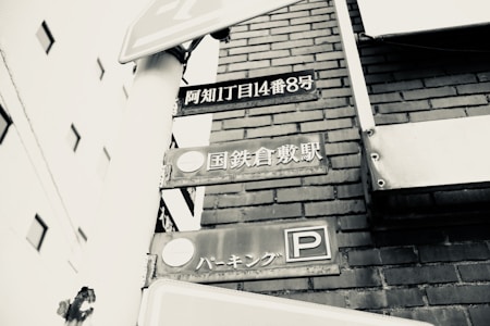 A black-and-white photograph featuring street signs with Japanese text mounted on a white post. The background includes a brick wall and parts of nearby buildings.
