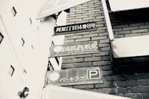 A black-and-white photograph featuring street signs with Japanese text mounted on a white post. The background includes a brick wall and parts of nearby buildings.