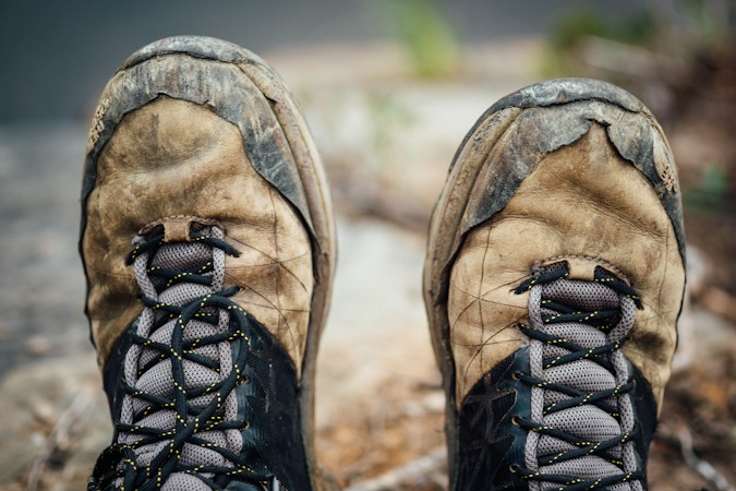 Close-up of two worn, mud-streaked tan suede and mesh hiking boots with gray laces on a rocky trail