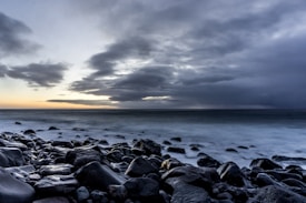 Dark, rocky shoreline meets a turbulent sea under a cloudy sky. The horizon is faintly illuminated by a gap in the clouds, suggesting a sunrise or sunset. The wet rocks in the foreground are smooth and reflect the muted light.
