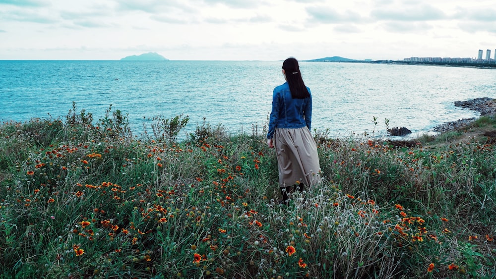 woman in blue blouse and gray skirt standing on red flower field beside body of water during daytime