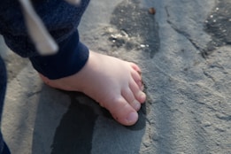 A small bare foot of a child is placed on a stone surface, with visible details of the toes and the smooth texture of the foot. The background shows the rough texture and natural color variations of the stone, with some moisture visible, likely from water.