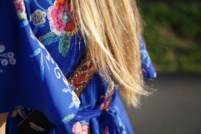 Long blonde hair drapes over a blue garment adorned with colorful embroidered flowers and patterns. The embroidered design features bright pink, red, green, and white threads, creating a vibrant contrast against the rich blue fabric. A black tag with text is partially visible on the lower left.