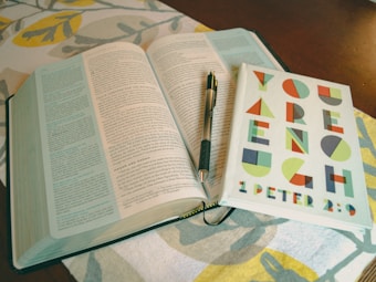 An open book with text in two columns lies on a cloth with a decorative leaf pattern. A notebook with colorful geometric letters spelling 'YOU ARE ENOUGH' and '1 PETER 2:9' is placed on top of the book, and a pen rests between the pages.