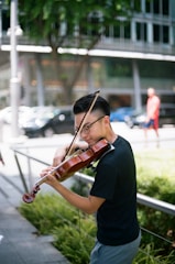 A person is playing a violin outdoors in an urban setting. The musician is focused and appears to be enjoying performing. In the background, there is greenery and a blurred view of a cityscape with modern buildings, cars, and a passerby.