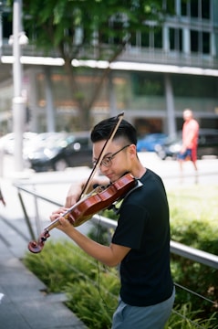 A person is playing a violin outdoors in an urban setting. The musician is focused and appears to be enjoying performing. In the background, there is greenery and a blurred view of a cityscape with modern buildings, cars, and a passerby.
