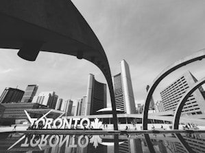 A black and white cityscape showcases modern architecture with curved arches framing a backdrop of tall buildings. The prominent 'Toronto' sign is visible near a reflective pool, adding to the urban atmosphere. People are seen walking around, contributing to a lively city vibe.