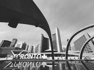 A black and white cityscape showcases modern architecture with curved arches framing a backdrop of tall buildings. The prominent 'Toronto' sign is visible near a reflective pool, adding to the urban atmosphere. People are seen walking around, contributing to a lively city vibe.