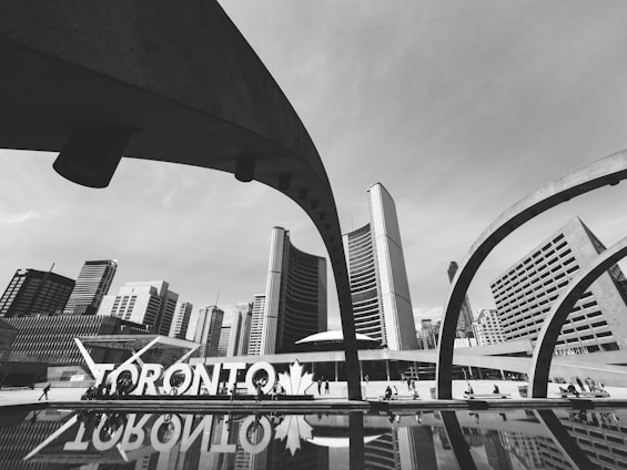 A black and white cityscape showcases modern architecture with curved arches framing a backdrop of tall buildings. The prominent 'Toronto' sign is visible near a reflective pool, adding to the urban atmosphere. People are seen walking around, contributing to a lively city vibe.