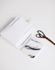 A neatly arranged set of office supplies including spiral notebooks, a binder clip, a pair of scissors, and a notebook with abstract art on the cover are placed on a clean white surface.