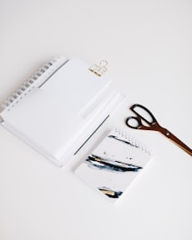 A neatly arranged set of office supplies including spiral notebooks, a binder clip, a pair of scissors, and a notebook with abstract art on the cover are placed on a clean white surface.
