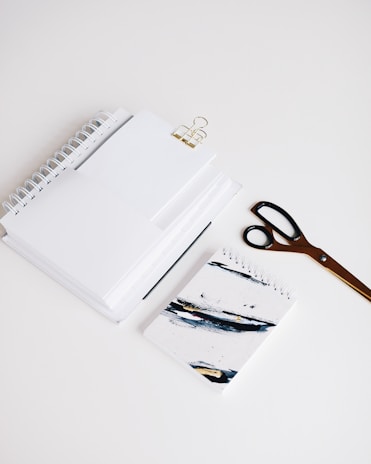 A neatly arranged set of office supplies including spiral notebooks, a binder clip, a pair of scissors, and a notebook with abstract art on the cover are placed on a clean white surface.