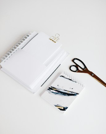 A neatly arranged set of office supplies including spiral notebooks, a binder clip, a pair of scissors, and a notebook with abstract art on the cover are placed on a clean white surface.