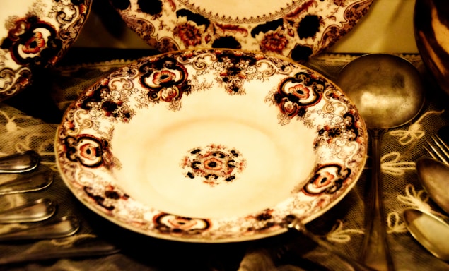 An intricately designed porcelain plate with ornate floral and geometric patterns, surrounded by antique silverware including spoons and a ladle on a lace tablecloth.