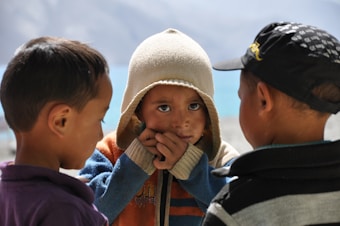 Three children are gathered closely together, with one child wearing a beige hooded sweater, looking directly at the camera with hands near the face. The child in the center is framed by two other children, one in a black cap and the other in a purple shirt, engaged in conversation or play.