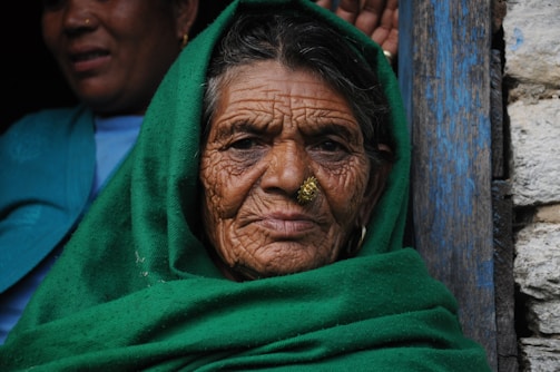 An elderly woman with deep wrinkles on her face is wearing a vibrant green shawl over her head. She has a gold nose ring and looks directly at the camera with a serene expression. Another person, partially visible, stands in the background wearing a blue garment.