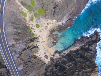 An aerial view captures a small secluded beach surrounded by rugged rocky cliffs and clear turquoise waters. People are seen relaxing on the sandy shore with colorful umbrellas and beach gear. The scene is bordered by a winding road to the left, lined with a guardrail.