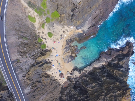 An aerial view captures a small secluded beach surrounded by rugged rocky cliffs and clear turquoise waters. People are seen relaxing on the sandy shore with colorful umbrellas and beach gear. The scene is bordered by a winding road to the left, lined with a guardrail.