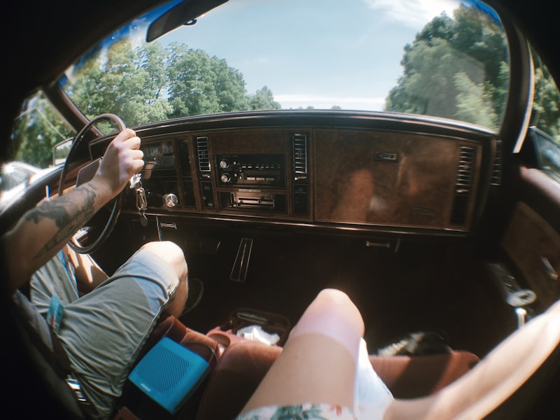Inside a car, the image captures the dashboard and steering wheel area. A person with tattooed arms is driving, wearing casual shorts. Their hand is on the steering wheel, and sunlight filters through the windshield. Another passenger's leg is partially visible, indicating a relaxed atmosphere. Outside, the scenery includes green trees under a clear blue sky.
