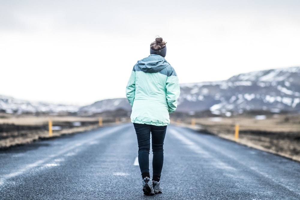 woman wearing teal jacket and black pants walking on concrete road