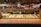 A display case filled with a variety of colorful pastries and cakes, including items topped with fruits and chocolate. Above the glass case, there are trays with croissants and other baked goods. The background shows people preparing food in a warm, inviting atmosphere with wooden shelves.