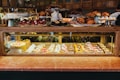 A display case filled with a variety of colorful pastries and cakes, including items topped with fruits and chocolate. Above the glass case, there are trays with croissants and other baked goods. The background shows people preparing food in a warm, inviting atmosphere with wooden shelves.