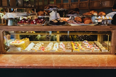 A display case filled with a variety of colorful pastries and cakes, including items topped with fruits and chocolate. Above the glass case, there are trays with croissants and other baked goods. The background shows people preparing food in a warm, inviting atmosphere with wooden shelves.