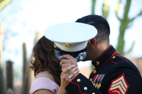 A couple is shown with the man wearing a formal military uniform decorated with medals and insignia, holding a white peaked cap. They are in an embrace, partially obscured by the cap, with cacti visible in the blurred background.