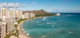 A coastal cityscape with high-rise buildings along a beach, surrounded by a turquoise ocean. In the background, a distinctive mountain range provides a stunning backdrop.