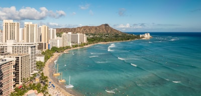 A coastal cityscape with high-rise buildings along a beach, surrounded by a turquoise ocean. In the background, a distinctive mountain range provides a stunning backdrop.