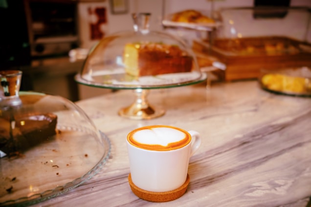 A marble countertop featuring a cup of cappuccino with a heart-shaped foam design, surrounded by various pastries displayed under glass domes. The background is softly blurred, highlighting the cup in the foreground.