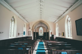 A serene, empty church interior with wooden pews on either side of a central aisle covered by a blue carpet. The architecture is traditional with arched windows along the light-colored walls, allowing natural light to stream in. Stained glass can be seen in the window near the altar, and a decorative ceiling adds to the peaceful atmosphere.