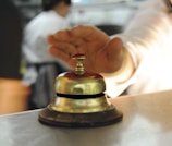 A hand is reaching towards a shiny, metallic desk bell set on a counter. The background is blurred, indicating a busy environment, likely in a reception or service area.