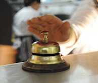 A hand is reaching towards a shiny, metallic desk bell set on a counter. The background is blurred, indicating a busy environment, likely in a reception or service area.