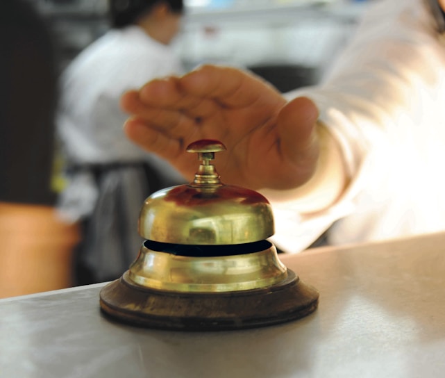 A hand is reaching towards a shiny, metallic desk bell set on a counter. The background is blurred, indicating a busy environment, likely in a reception or service area.
