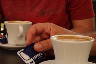 A close-up of two white cups of coffee sits on saucers on a black table. A person in a red shirt holds a blue and white sugar packet labeled 'CAFFECEL espresso' with one hand while the other hand cups one of the coffee mugs.