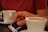 A close-up of two white cups of coffee sits on saucers on a black table. A person in a red shirt holds a blue and white sugar packet labeled 'CAFFECEL espresso' with one hand while the other hand cups one of the coffee mugs.